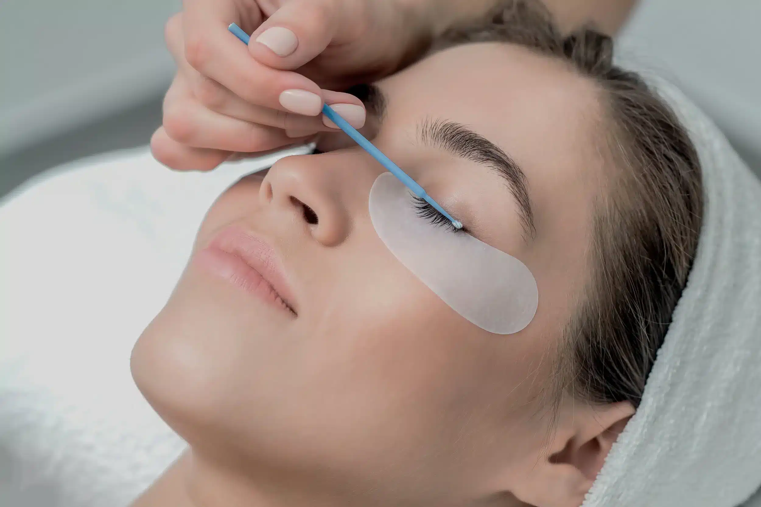 A woman lying down with her eyes closed, receiving an eyelash cleaning or treatment using a blue applicator, with a protective pad under her eye.