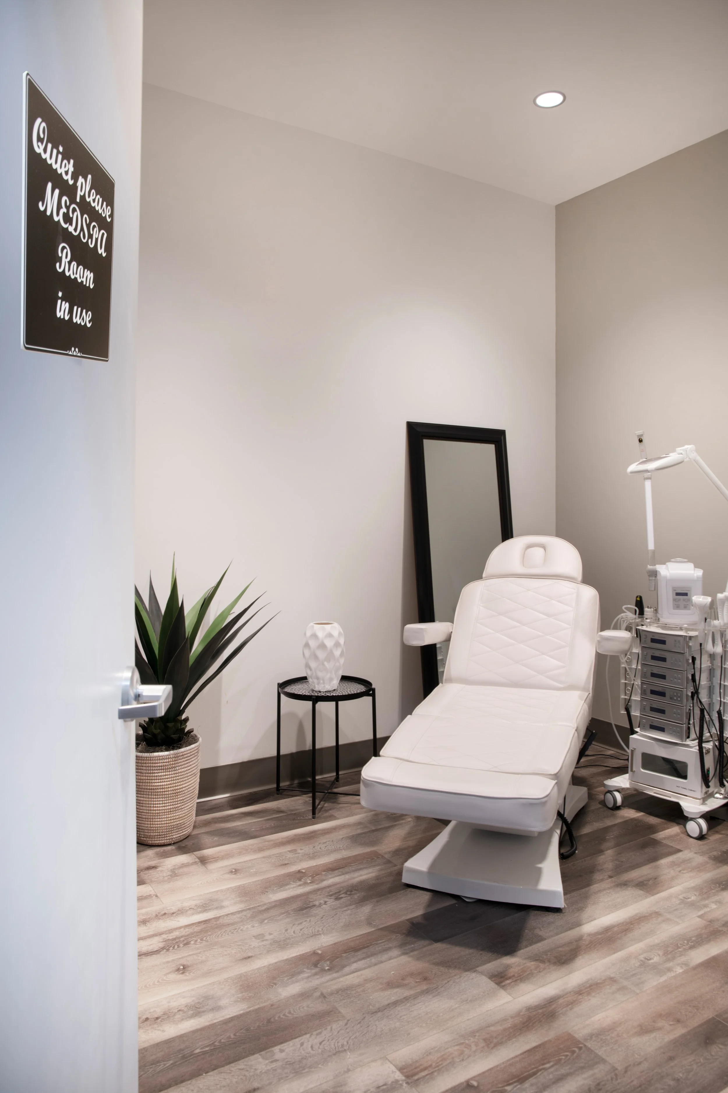 A medical examination room with a white reclining chair, a standing mirror, a small black side table with a white textured vase, a potted plant, and medical equipment on a mobile cart. There is a sign on the wall that says "Quiet please MECSOA Room in use."